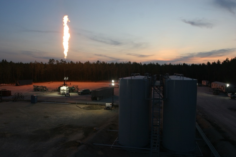 Gas flare illuminates the dusk sky at a well pad with storage tanks and field equipment